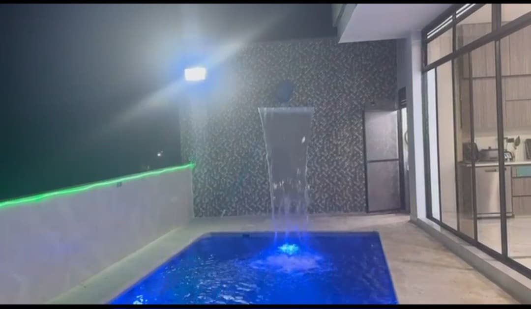 A modern outdoor space features a small illuminated pool with a waterfall and vibrant blue lighting. The area is surrounded by a textured wall and has glass doors that offer a glimpse into the interior, creating a harmonious connection between indoor and outdoor environments.