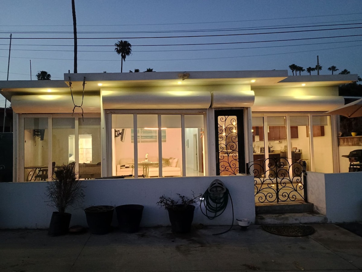 The exterior of the beachfront home is showcased at dusk, with warm light emanating from the large glass doors and windows. The entrance features an ornate wrought iron door, and potted plants decorate the area. The open layout allows for a glimpse of the living space and kitchen inside.