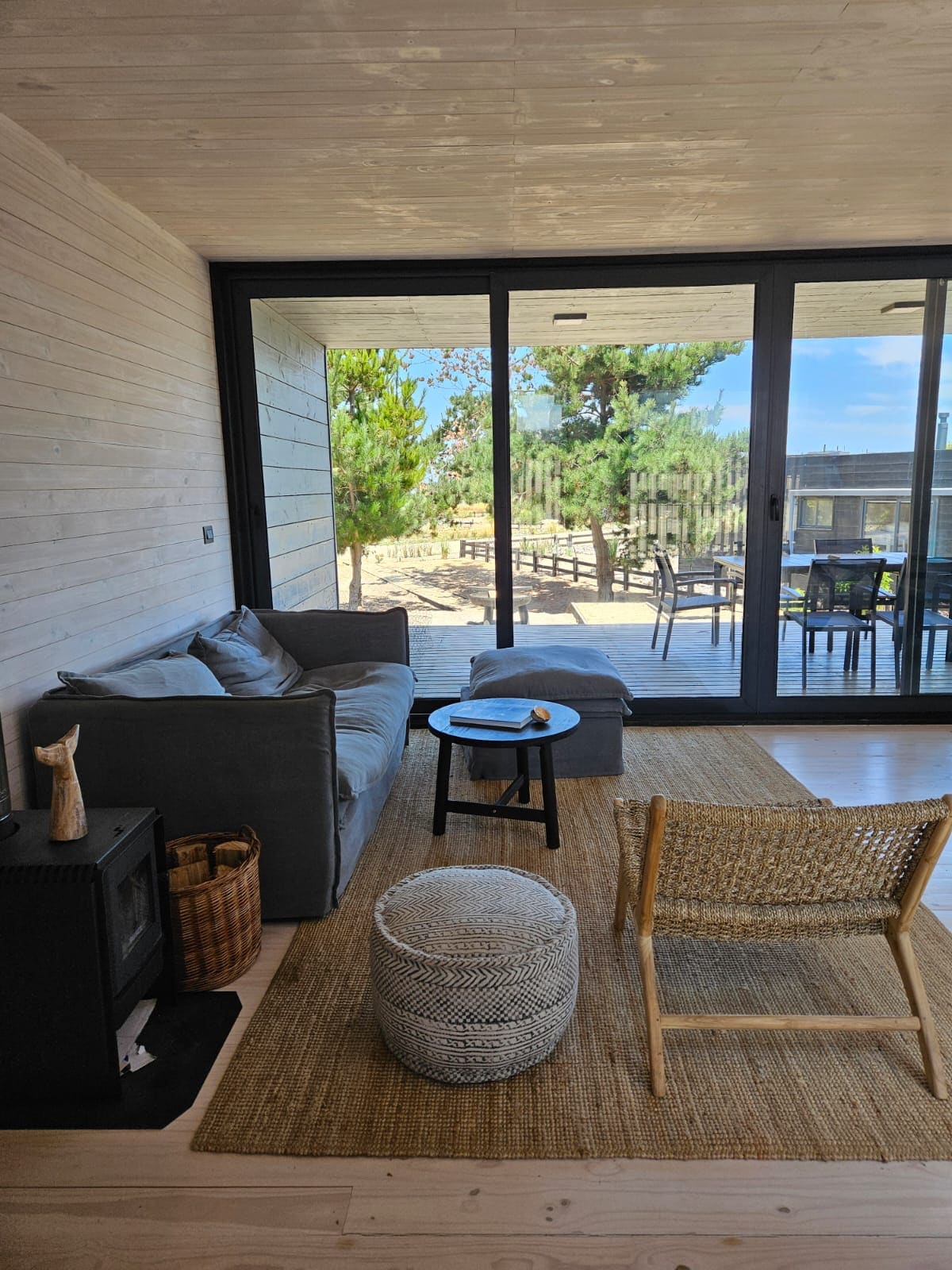 A cozy living space features a comfortable gray sofa positioned against a natural wood wall. A small round table is placed nearby, with light streaming through expansive glass doors that open to a terrace. A woven chair and basket add texture to the room.