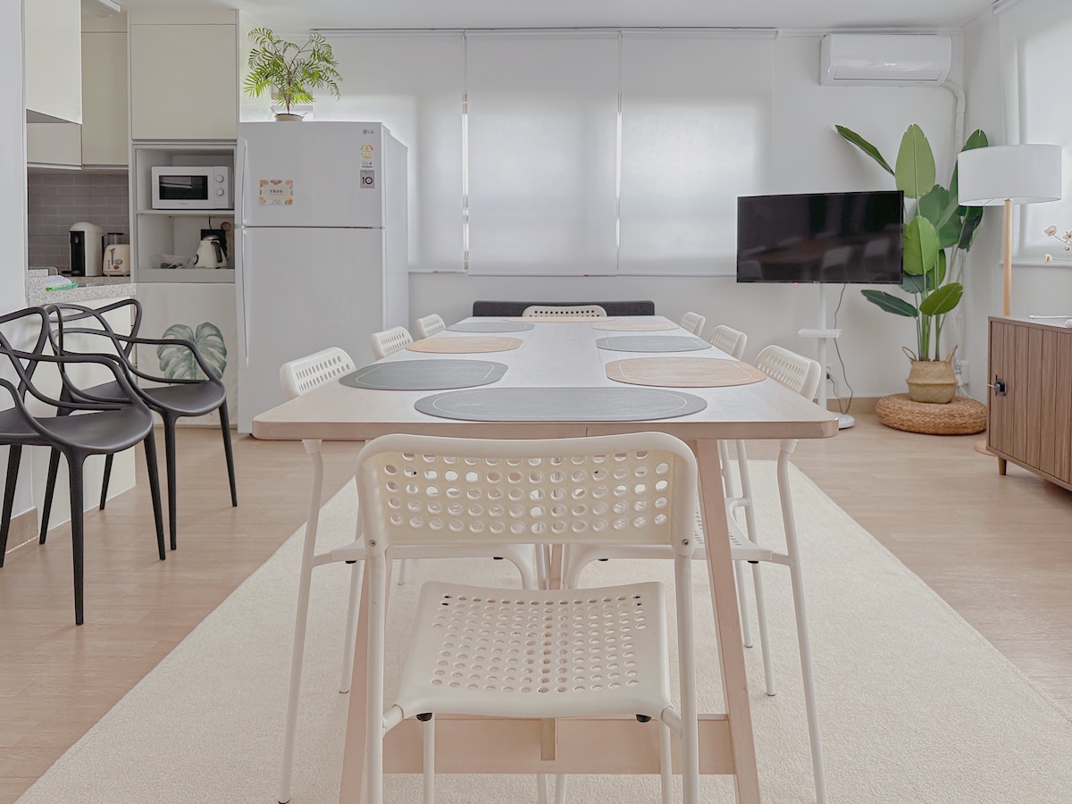 A spacious dining area features a long table surrounded by stylish white chairs. A large window allows natural light to fill the room, while a leafy plant adds a touch of greenery. A comfortable couch and a television are visible in the backdrop.