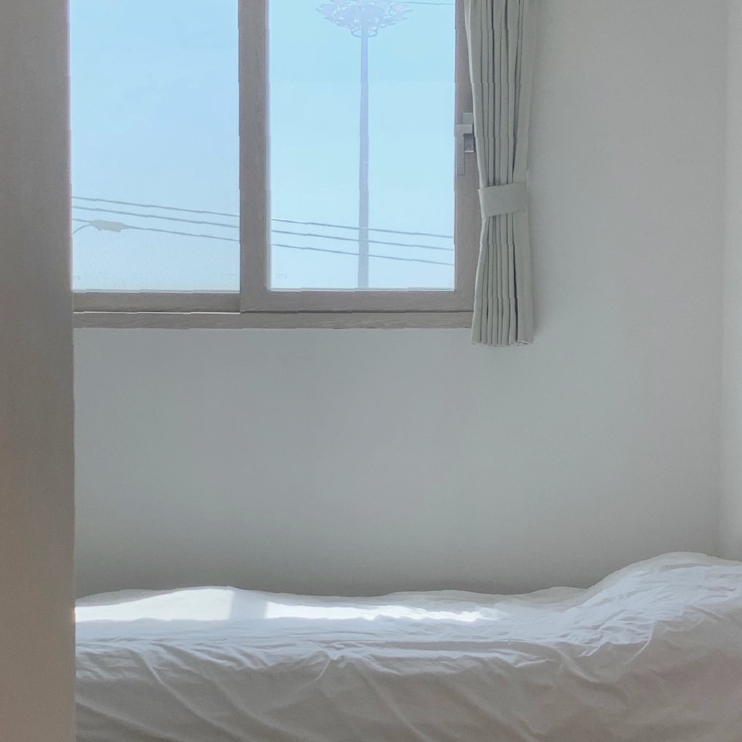 A bright sleeping area is shown, featuring a spacious bed with a plain white comforter. Natural light enters through a window fitted with light curtains. Outside, a street lamp is visible against a clear sky.