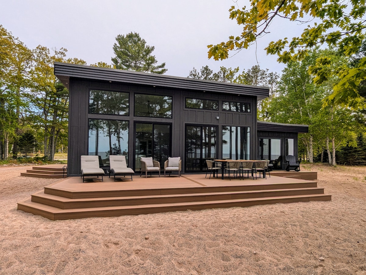 The modern exterior of a black house features large floor-to-ceiling windows, providing expansive views. A spacious deck is adorned with comfortable lounge chairs and a dining table, all set within a landscaped sandy area surrounded by trees.