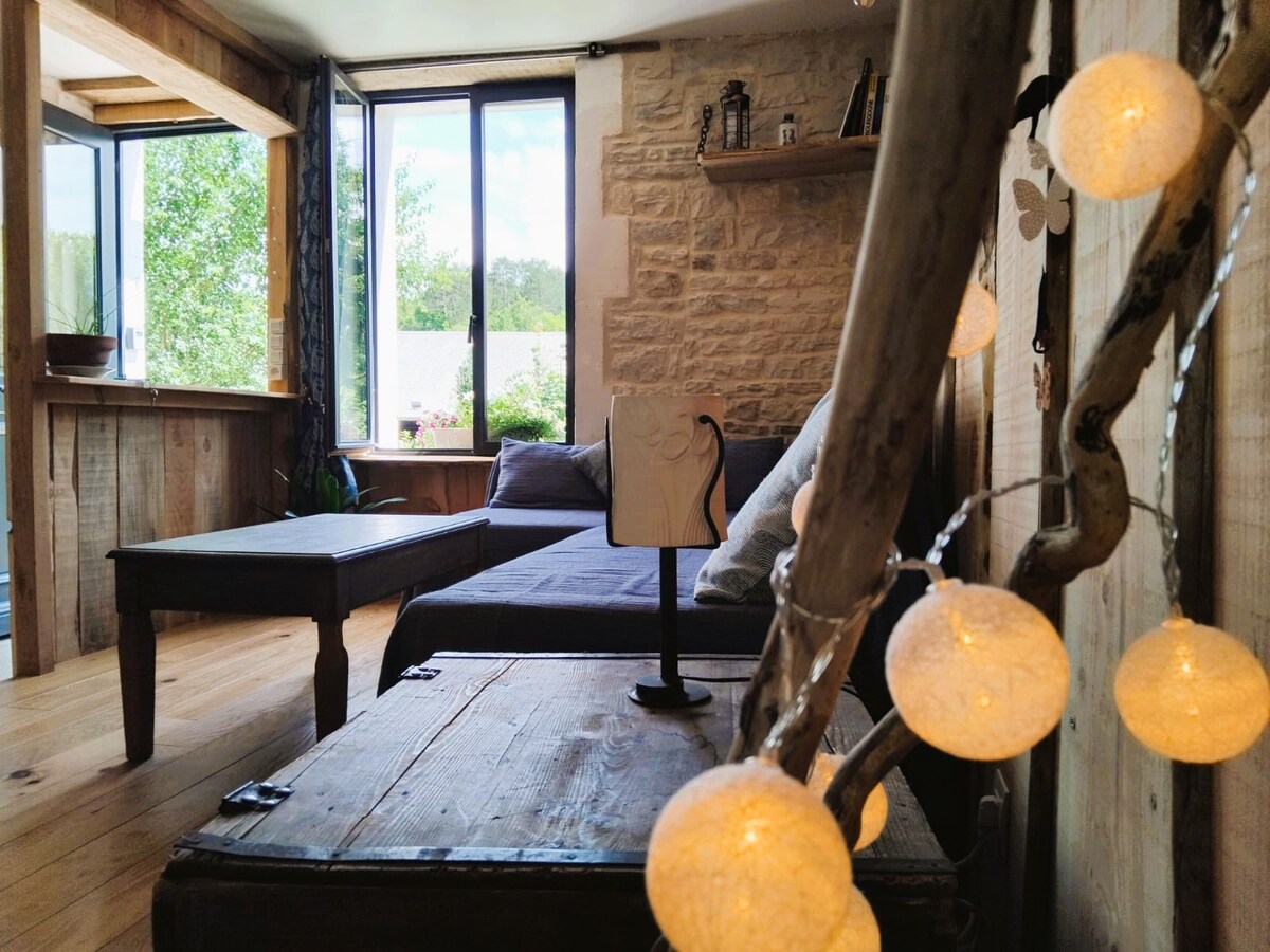 A warm living area features a comfortable sofa and a wooden coffee table. Natural light enters through large windows, illuminating the rustic stone walls. Decorative string lights add a cozy charm, while a small shelf holds personal touches against a backdrop of natural textures.