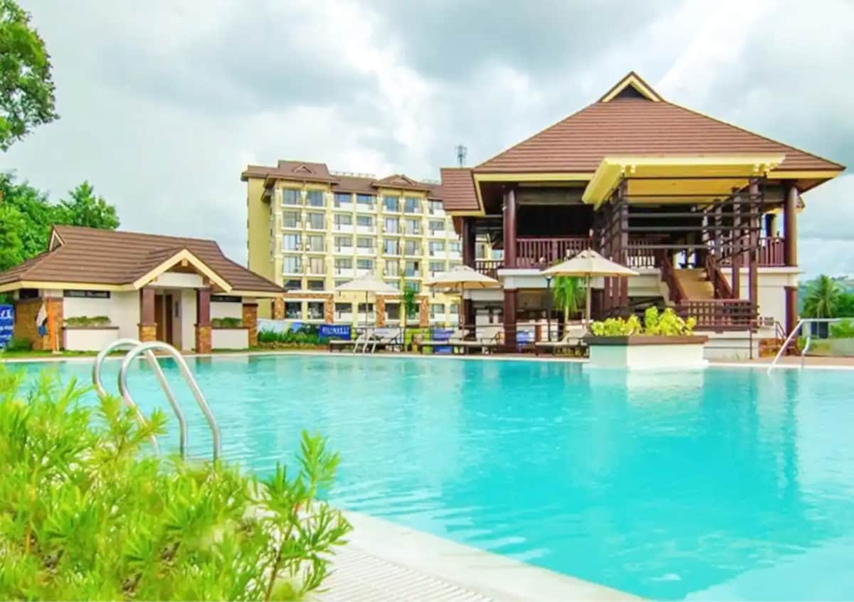 A large outdoor pool is framed by lush greenery, with lounge chairs and umbrellas positioned around its perimeter. A two-story building with a classic architectural style is visible in the background, featuring balconies and a welcoming entrance.