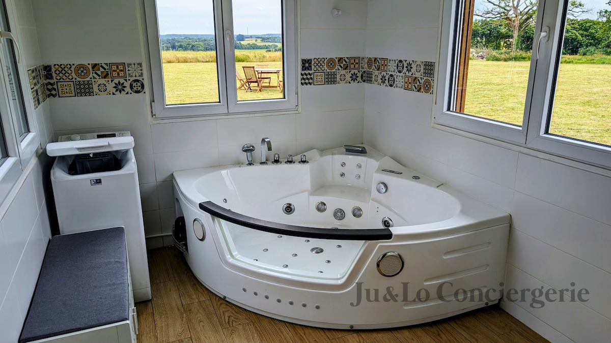 A spacious bathroom features a modern jacuzzi tub positioned under large windows, offering views of the surrounding nature. A washer can be seen beside the tub, with a minimalist bench nearby. Decorative tiles line the walls, complementing the serene environment.