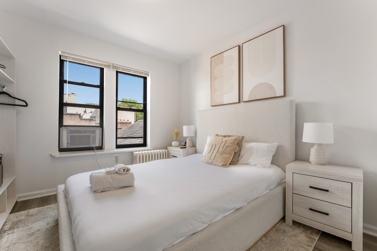 A bright and airy bedroom features a plush sleeping area with a comfortable bed adorned with soft linens. Two windows allow natural light to fill the space, while modern artwork hangs above the headboard. A nightstand with a lamp and a beige accent pillow enhances the decor.