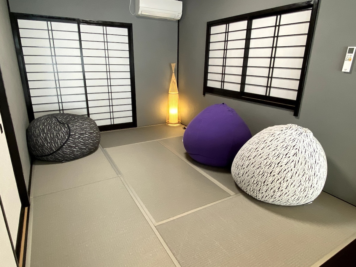 A relaxing space features three large bean bags in different colors, arranged on a tatami mat floor. Natural light filters through shoji screens, creating a serene atmosphere. A warm light source stands nearby, adding a gentle glow to the room.