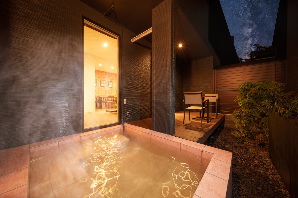 An outdoor hot spring bath is shown in a serene setting, with steam rising gently above the warm water. A glass door leads to a cozy seating area, while the evening sky is visible filled with stars, enhancing the tranquil environment.