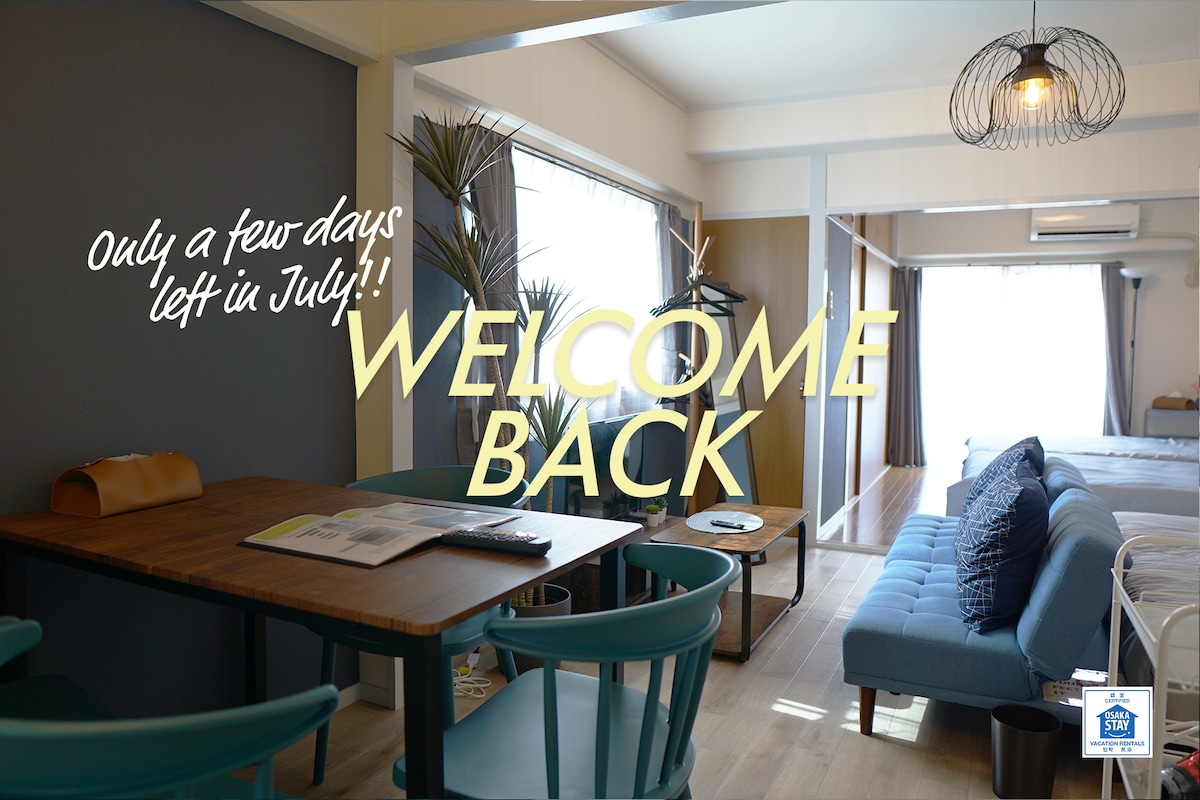 A well-furnished living area features a dining table with chairs and a blue sofa. Bright natural light enters through large windows, creating a fresh atmosphere. Decorative plants add a touch of greenery, while the cozy layout accommodates various seating options for relaxation.