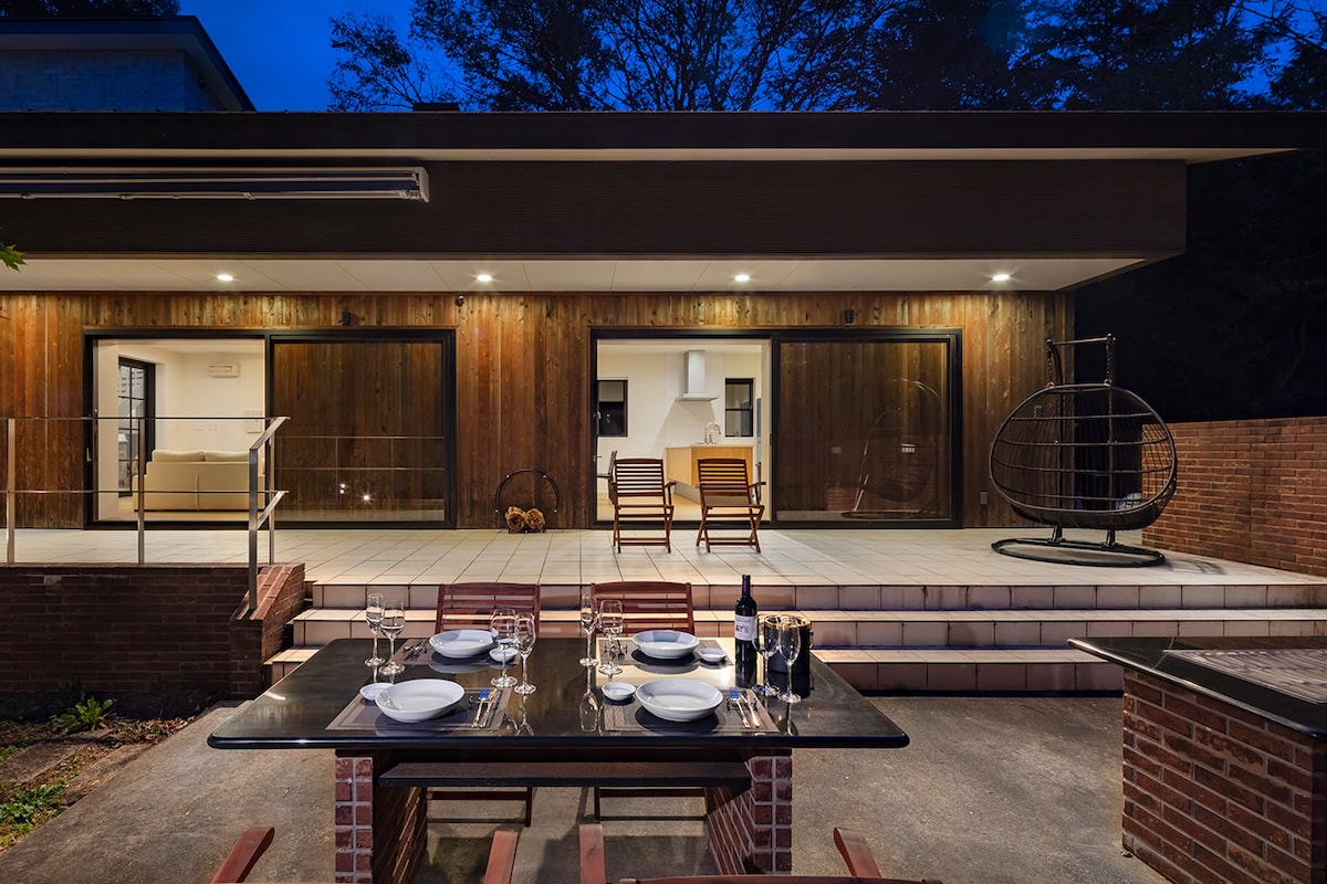 An outdoor dining area is set with a table, plates, and glasses, positioned on a stone patio. The back of the building features large glass doors that open to a spacious interior, with a warm wooden facade visible in the evening light.