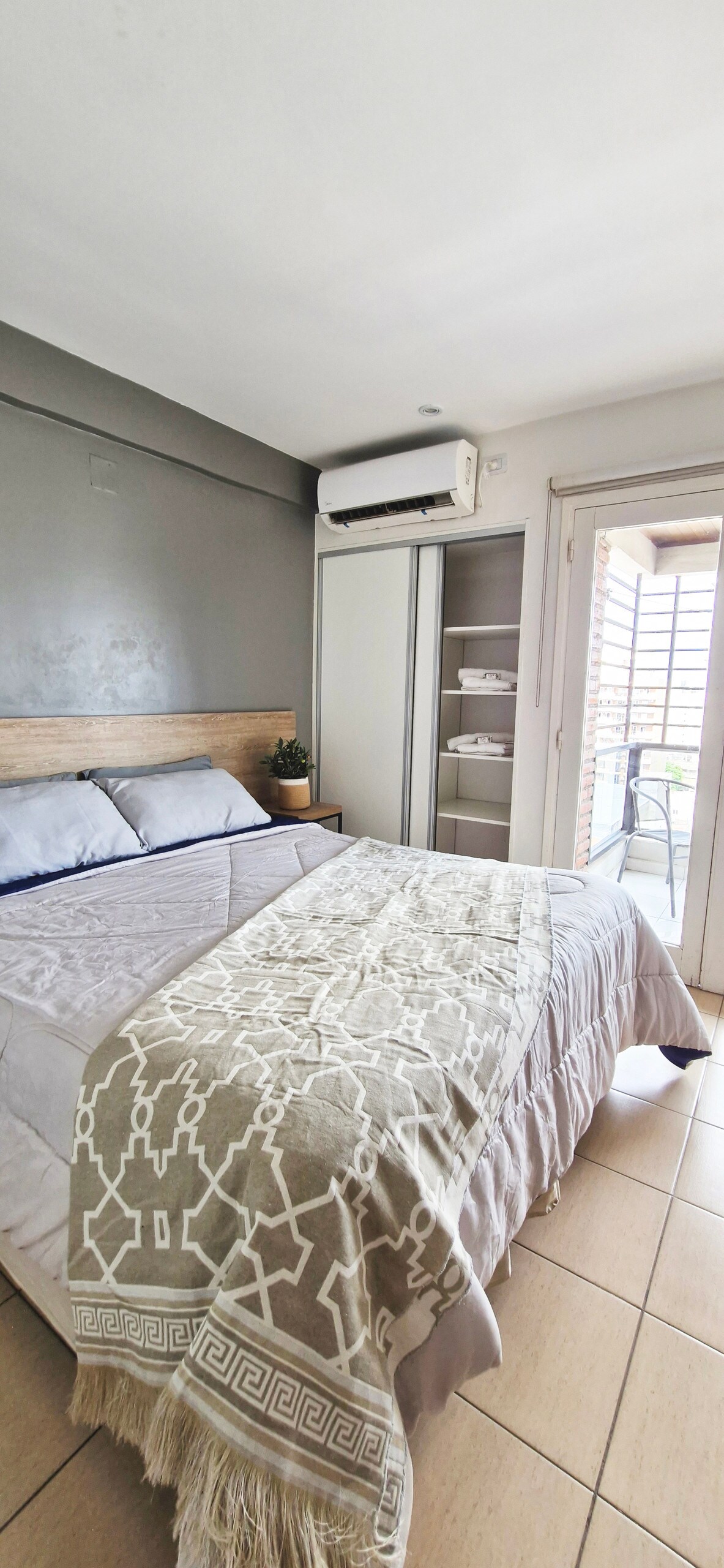 A spacious bedroom is presented with a comfortable sleeping arrangement featuring a light blanket draped over the bed. Natural light filters through the sliding door to the balcony, which offers an outdoor view. A wardrobe with neatly arranged linens is visible beside the bed.