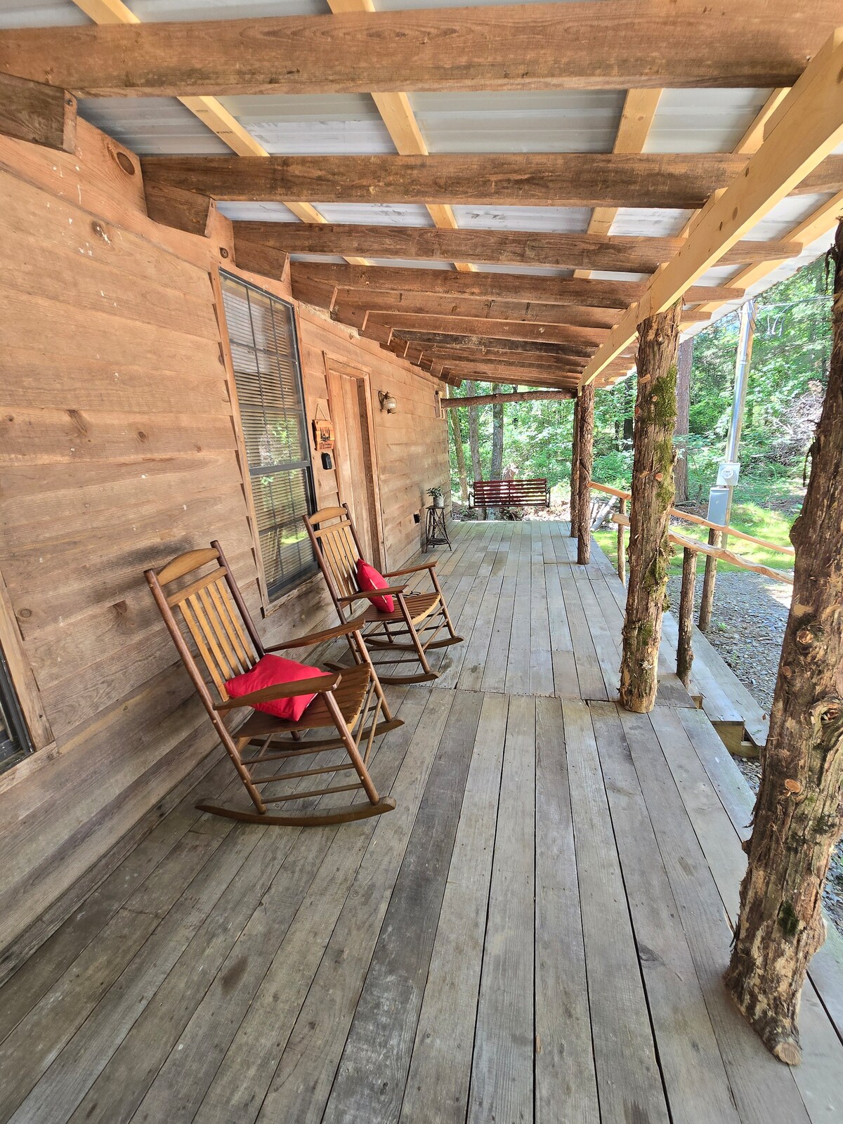 A welcoming porch extends along the cabin, featuring two wooden rocking chairs with red cushions. Natural wood beams support the roof, while the floor is composed of spacious wooden planks. Surrounding greenery is visible, enhancing the connection to the wooded setting.