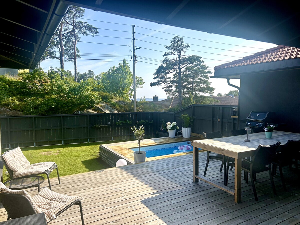 A wooden deck area features a dining table surrounded by chairs, with a gas grill positioned nearby. A pool can be seen set within the deck. Lush greenery and trees provide a natural backdrop, creating a serene outdoor environment.