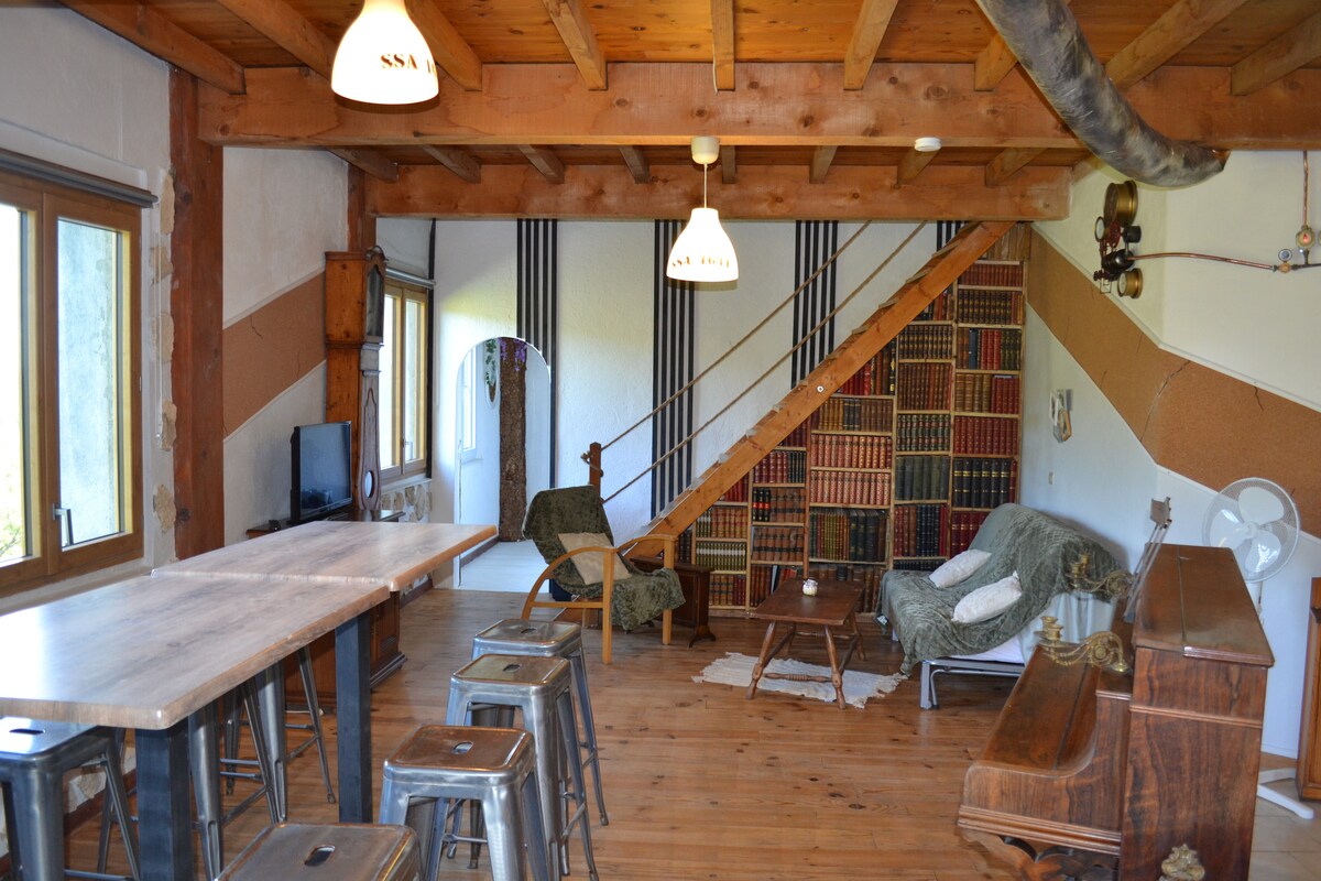 An open living area features wooden beams and a staircase leading to an upper level. A large table with stools occupies one side, while a cozy seating arrangement includes a sofa draped in a blanket. Natural light floods in through spacious windows, enhancing the inviting atmosphere.