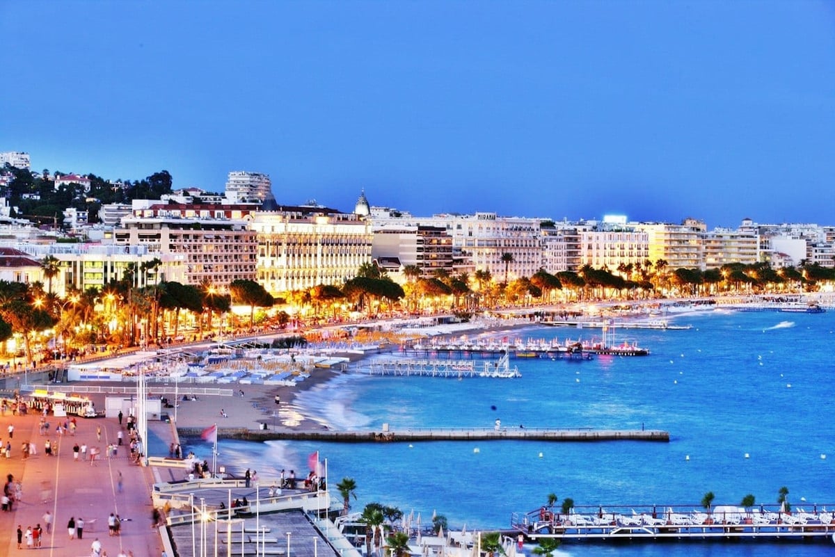 The scene captures a lively beachfront promenade at twilight, lined with illuminated buildings and palm trees. The gentle waves of the sea reflect the soft lights from surrounding establishments, creating a serene environment for evening strolls. Various individuals can be seen enjoying the promenade.