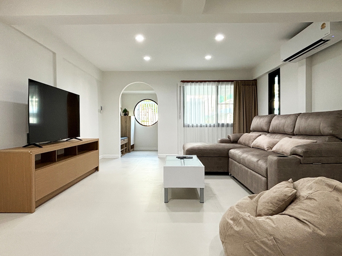 The bright living area features a comfortable sectional sofa and a large television, positioned across from a simple coffee table. Natural light enters through a large window, complemented by soft curtains. A cozy bean bag and an arched entryway create an inviting atmosphere.