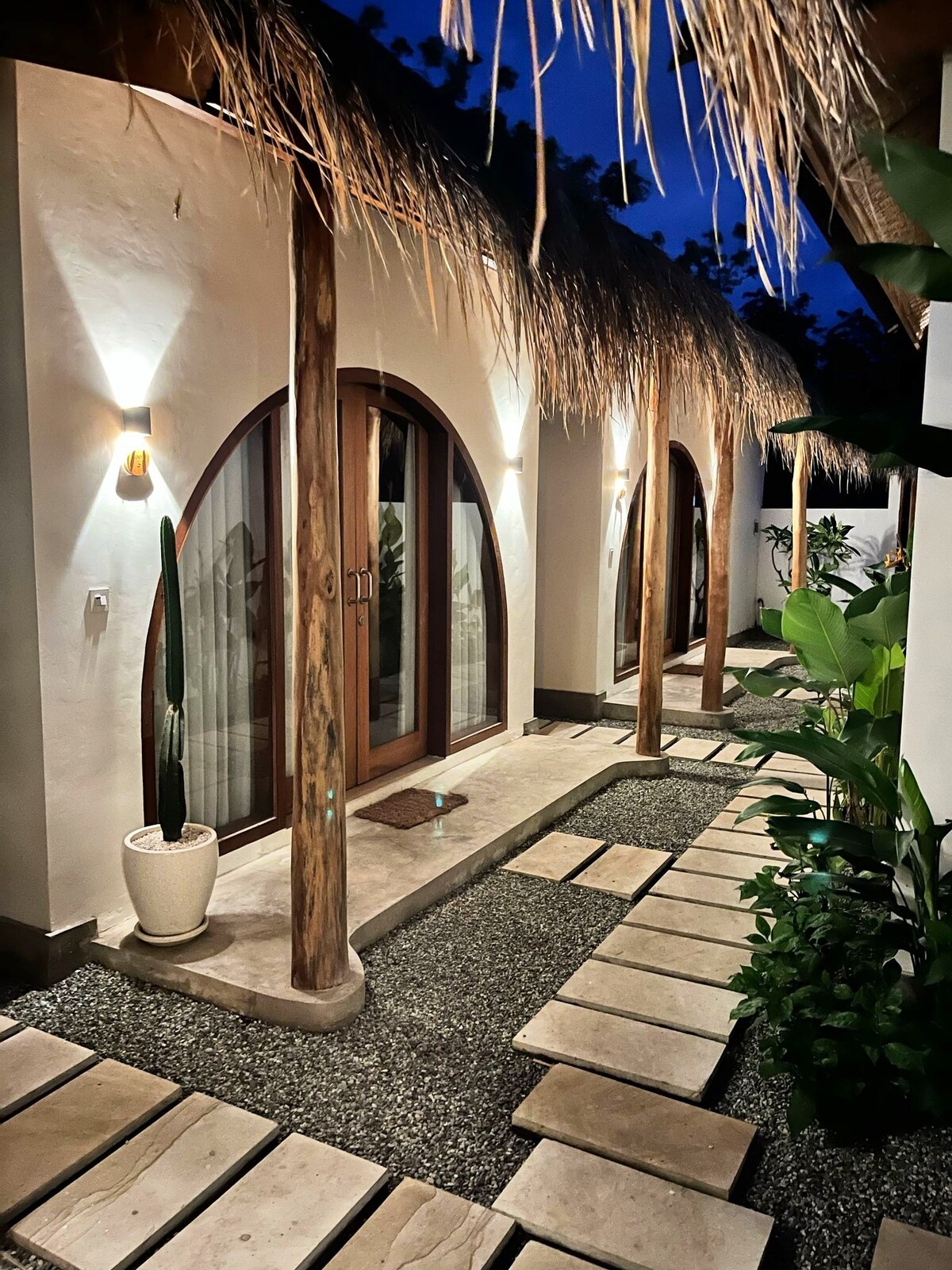 A serene pathway is lined with stone tiles, leading to cottage entrances adorned with arched wooden doors. Soft lighting from wall sconces creates a welcoming ambiance, while tropical plants and a decorative cactus add natural beauty along the walkway.