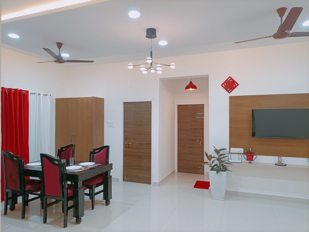 The dining area features a wooden table surrounded by four red-upholstered chairs. A sleek wall-mounted TV is visible alongside a decorative plant. Two wooden doors lead to private spaces, and overhead lighting enhances the modern decor. White walls and polished tiles create a bright atmosphere.