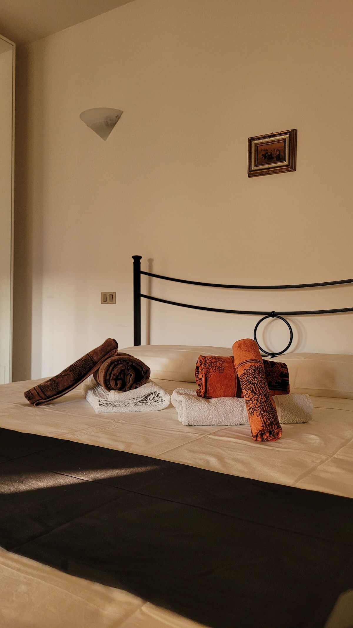 A spacious bed is adorned with neatly arranged towels in various colors. A small framed picture hangs above the bed, and soft light illuminates the room, highlighting the minimalist decor and the contrasting black bed covering.