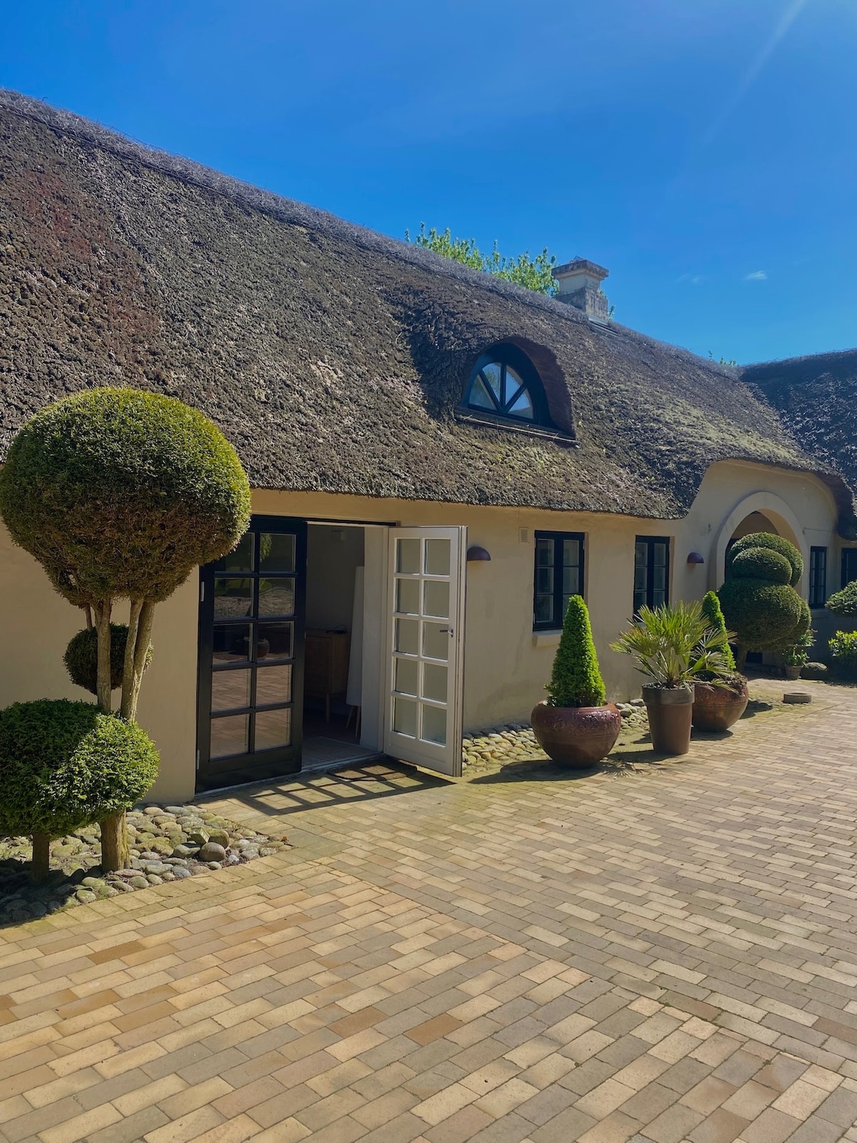 A charming thatched-roof cottage is framed by well-manicured shrubbery and vibrant potted plants. The entryway is highlighted by a dark wooden door and an arched window above, inviting guests into a serene outdoor space lined with brick paving.