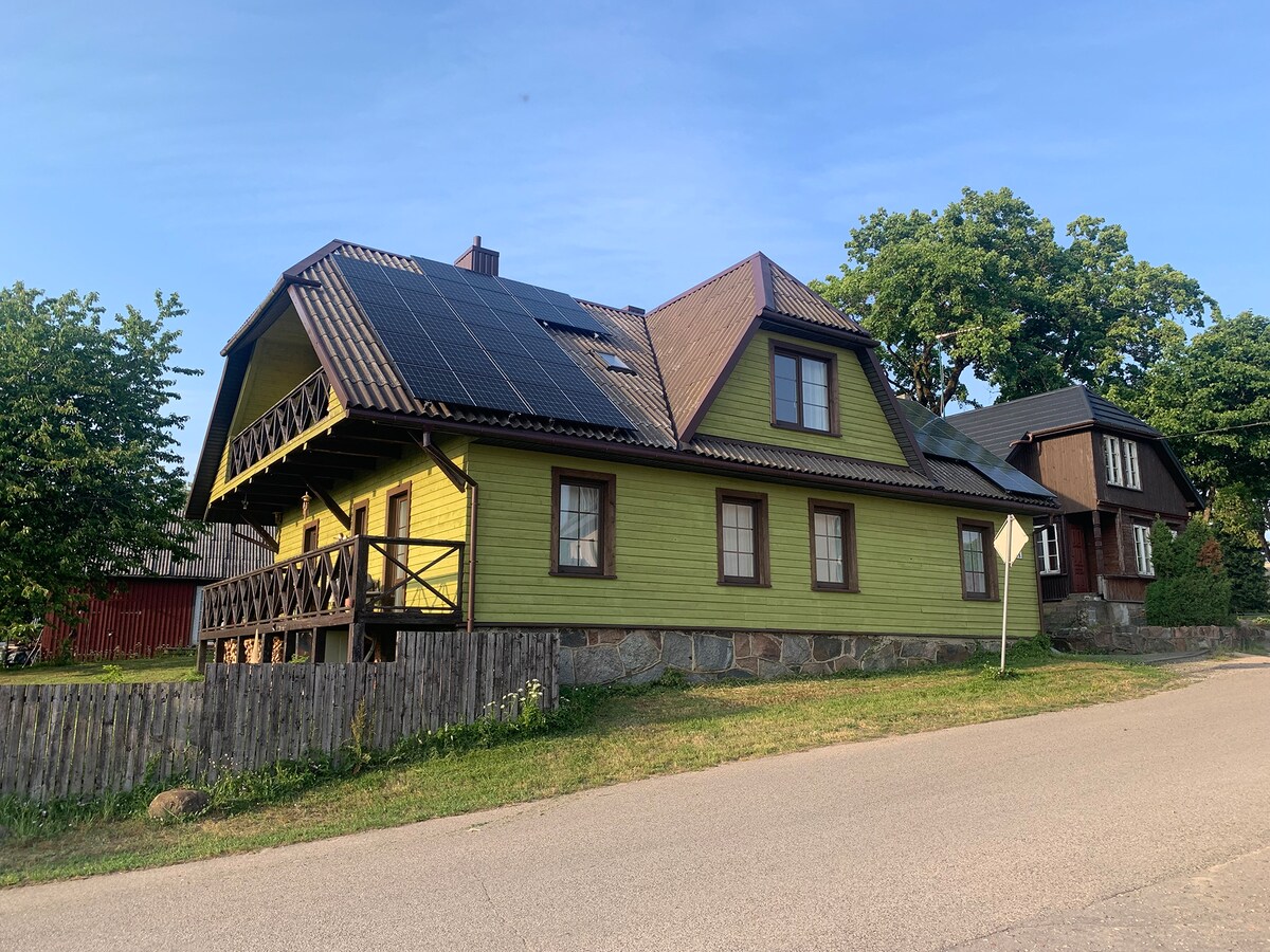 A green wooden house with a sloped roof is shown, featuring solar panels and a wraparound porch. The exterior is complemented by stone accents and surrounded by trees. A neighboring building with a darker exterior can be seen in the background.