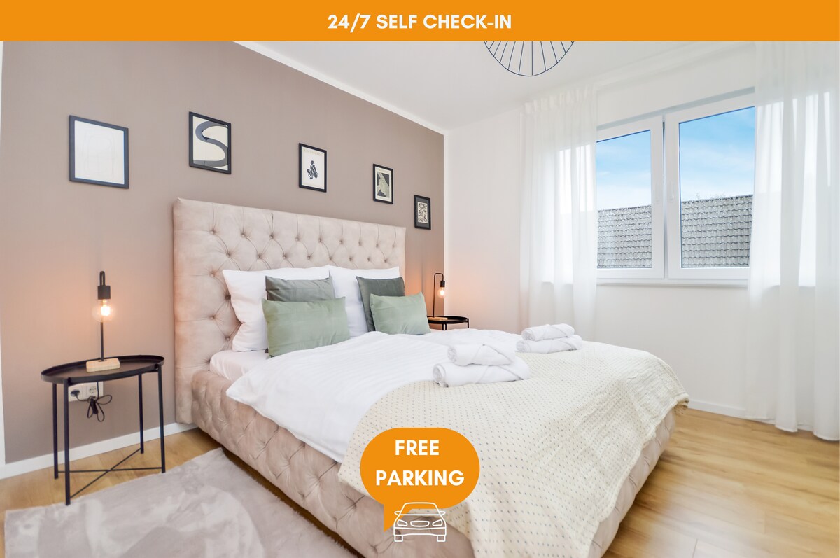 A spacious bedroom features a large tufted bed adorned with soft pillows and a light blanket. Soft natural light enters through the window, illuminating the room, while stylish wall art and nightstands enhance the décor. Ample space surrounds the cozy sleeping area.
