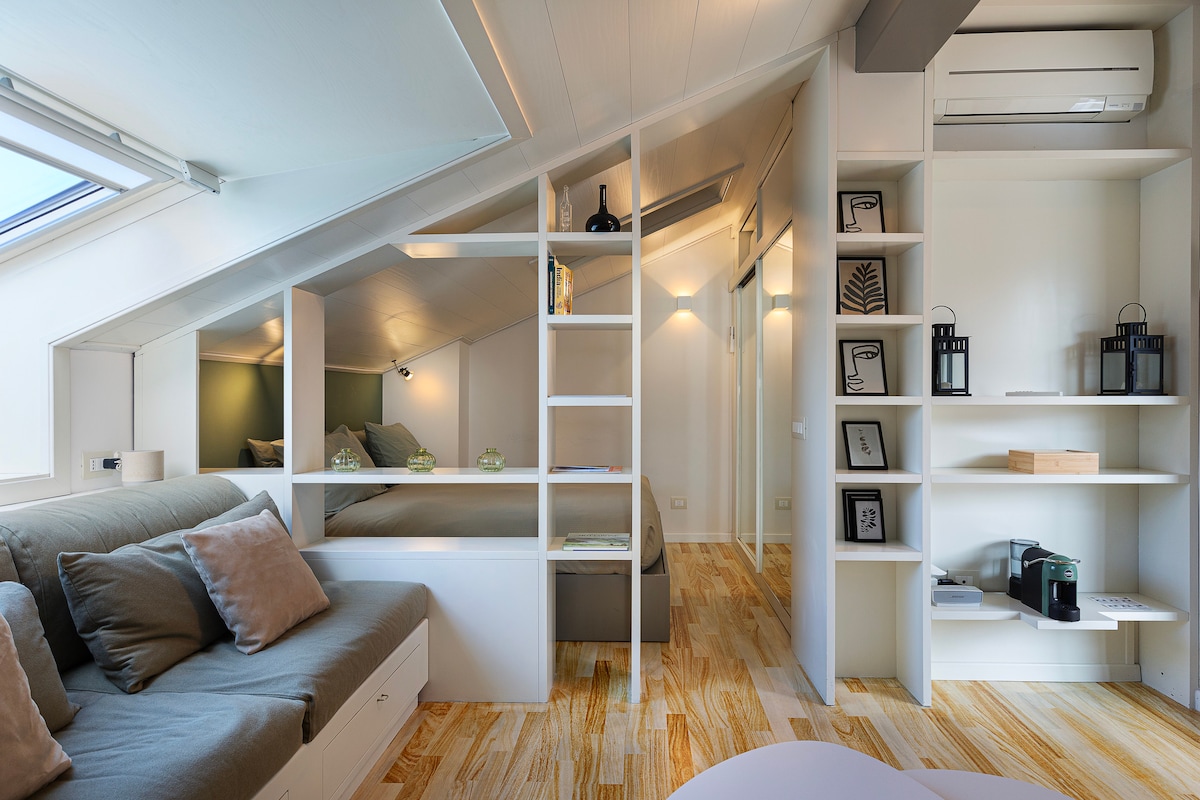 The open living space features a cozy seating area with soft cushions and a coffee table. Built-in shelving units create distinct zones, showcasing decorative items and practical appliances. Natural light filters through windows, illuminating the room while subtle ambient lighting highlights the wooden floor.