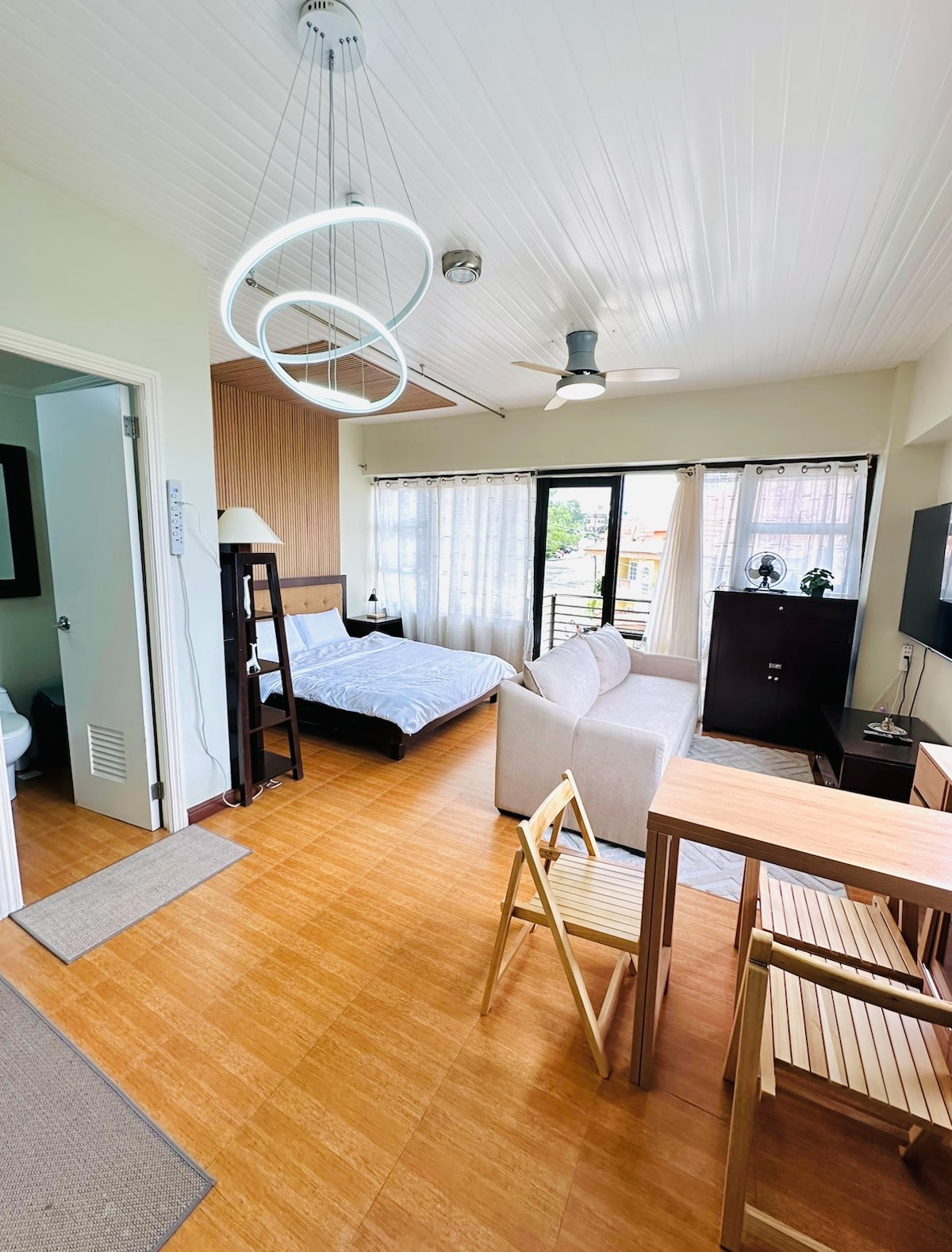 A spacious studio unit features a well-lit area with large windows allowing natural light. A cozy bed is complemented by a neutral-toned sofa and a dining area with wooden chairs. A modern light fixture hangs from the ceiling, creating an inviting space.