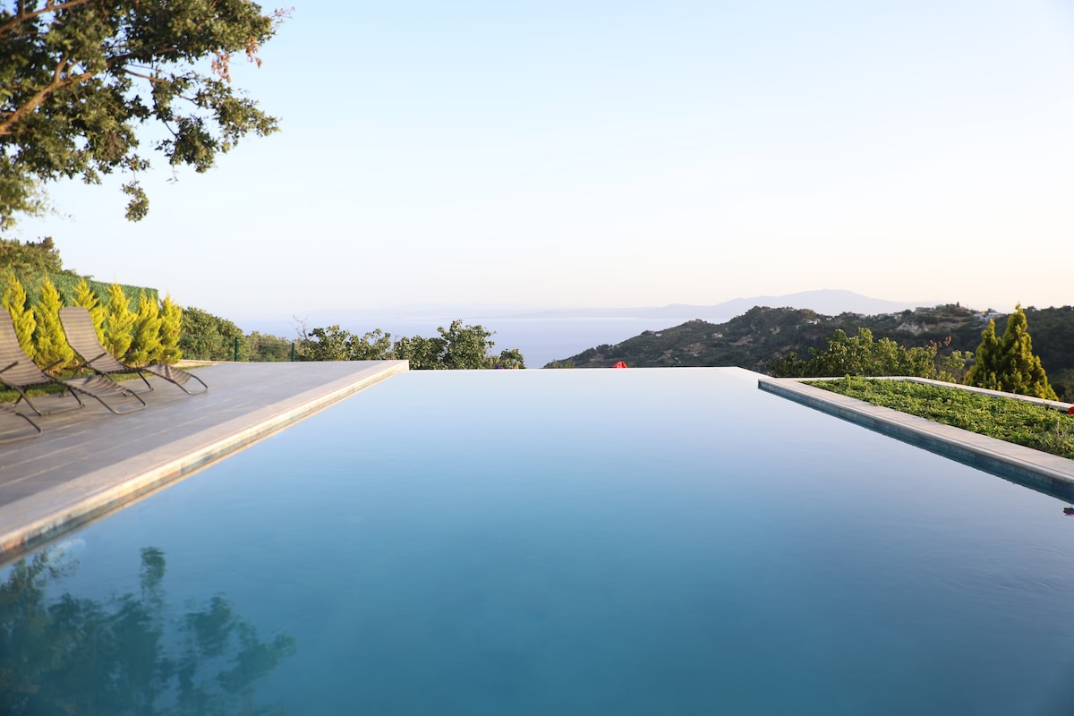 An infinity pool reflects the sky, seamlessly extending towards the horizon. Lush greenery surrounds the area, and lounge chairs are positioned nearby, offering a place to relax while enjoying the expansive views of the hills and distant sea.