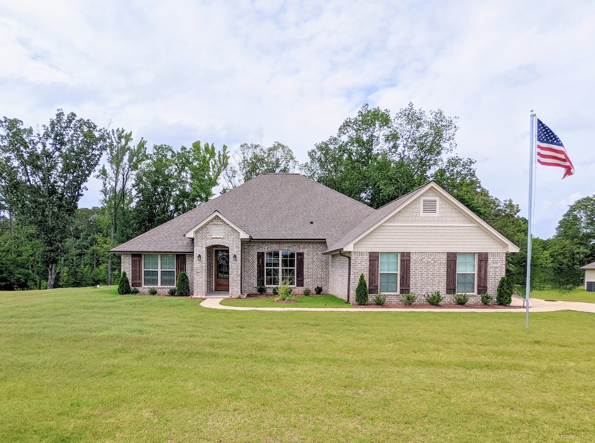 The front exterior of a single-story brick home is presented, featuring a well-maintained lawn with green grass and landscaped bushes. An American flag is displayed on a flagpole, while surrounding trees are visible in the background under a partly cloudy sky.
