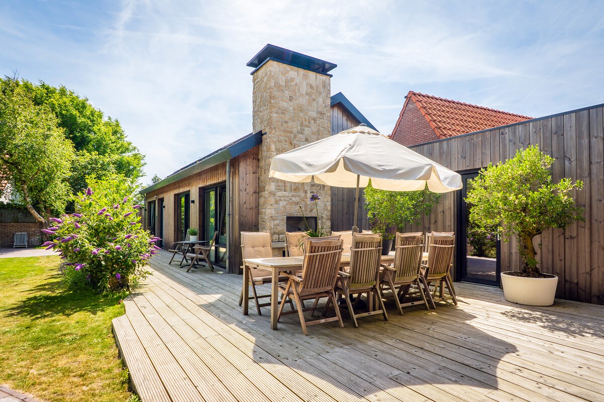 An outdoor patio area is visible, featuring a wooden deck with a large table surrounded by several chairs. A spacious umbrella provides shade over the seating. Lush greenery and flowering plants enhance the space, complemented by a stone chimney structure in the background.