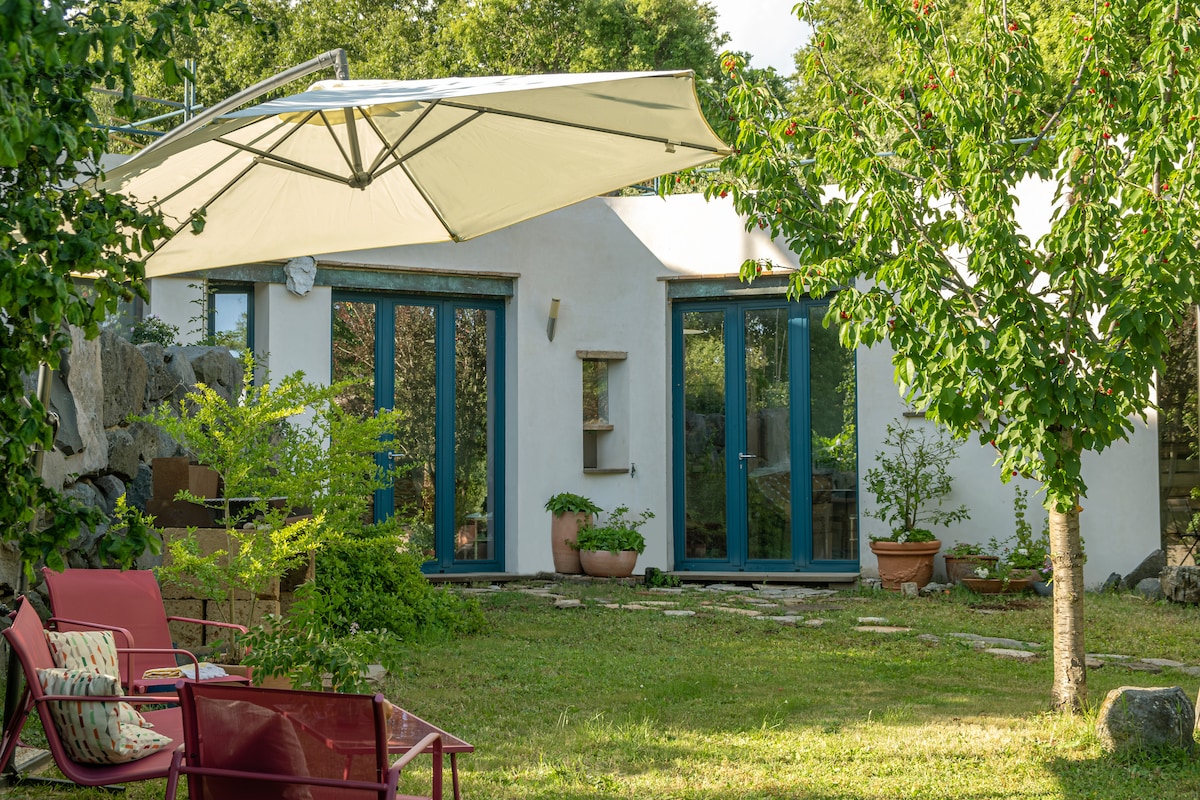 A well-maintained garden features a green lawn framed by shrubs and flowering plants. Two sets of blue doors lead into the building, while a large umbrella provides shade over a seating area with red chairs, inviting relaxation outdoors.