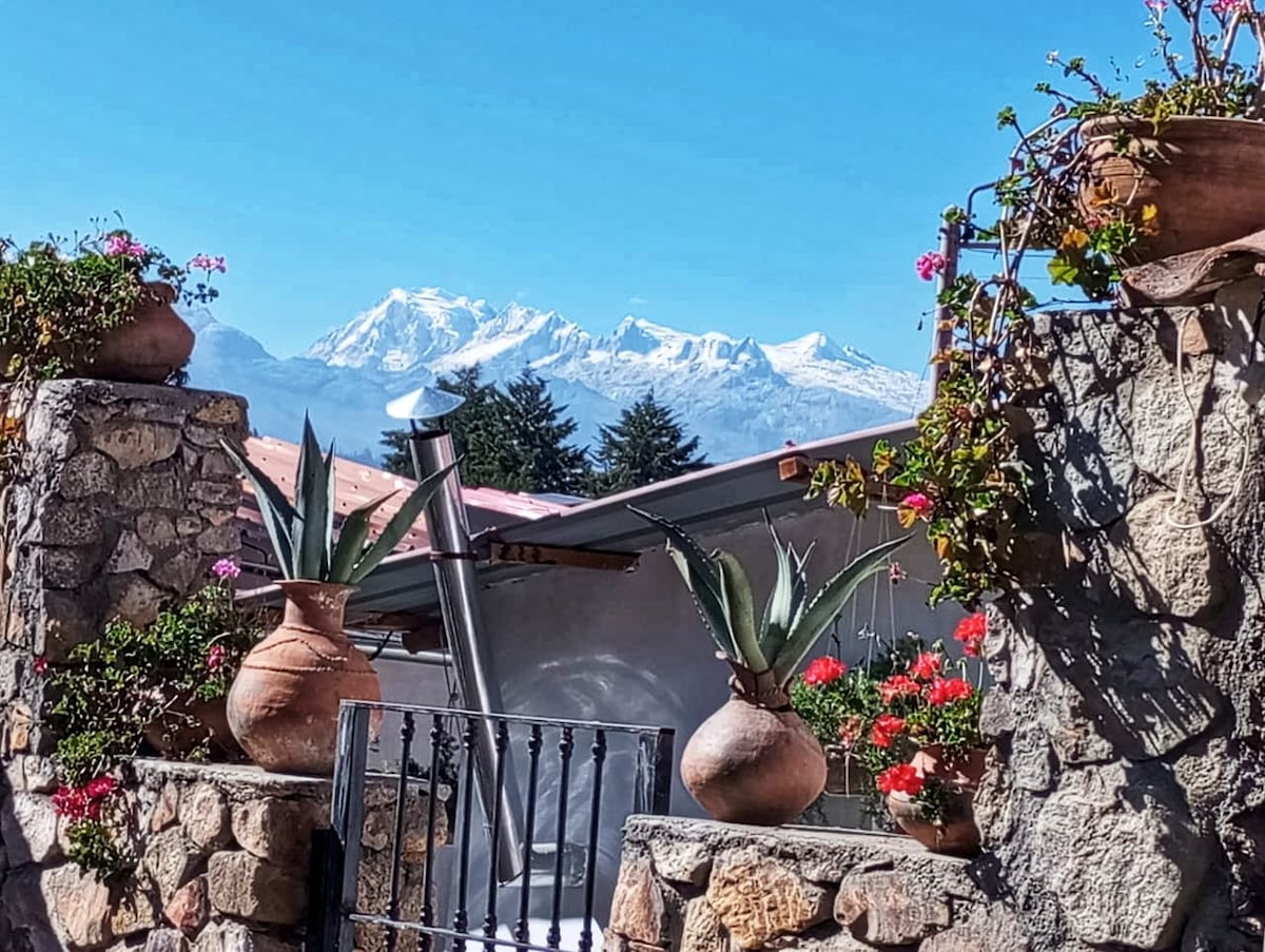 A stone wall adorned with potted plants frames a scenic view of snow-capped mountains in the distance. Colorful flowers in terracotta pots add vibrancy, while a simple metal gate offers access to the property. A clear blue sky complements the serene atmosphere.