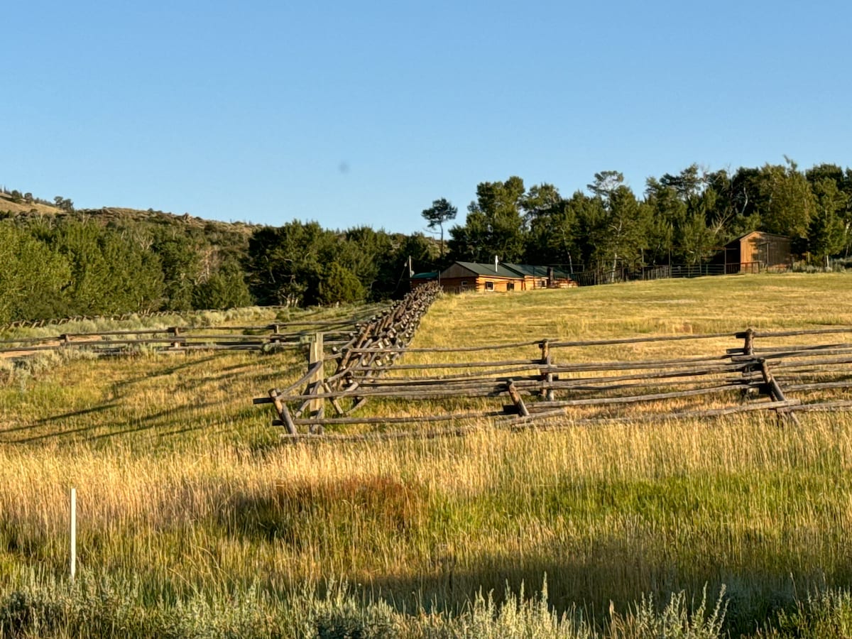 The cabin is situated at the end of a grassy field, framed by wooden fencing. Surrounding trees provide a natural backdrop against the clear blue sky. The serene landscape is highlighted by gentle slopes leading up towards the mountain.