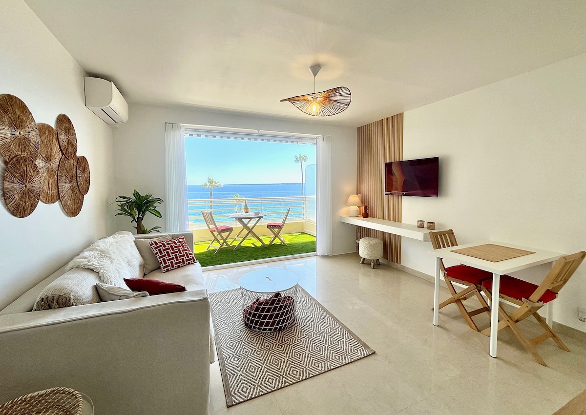 A bright living space is featured, with expansive windows offering a view of the sea. A comfortable sofa and a small dining table are situated against the wall. Decorative elements include a textured area rug and wall art, creating a serene environment.