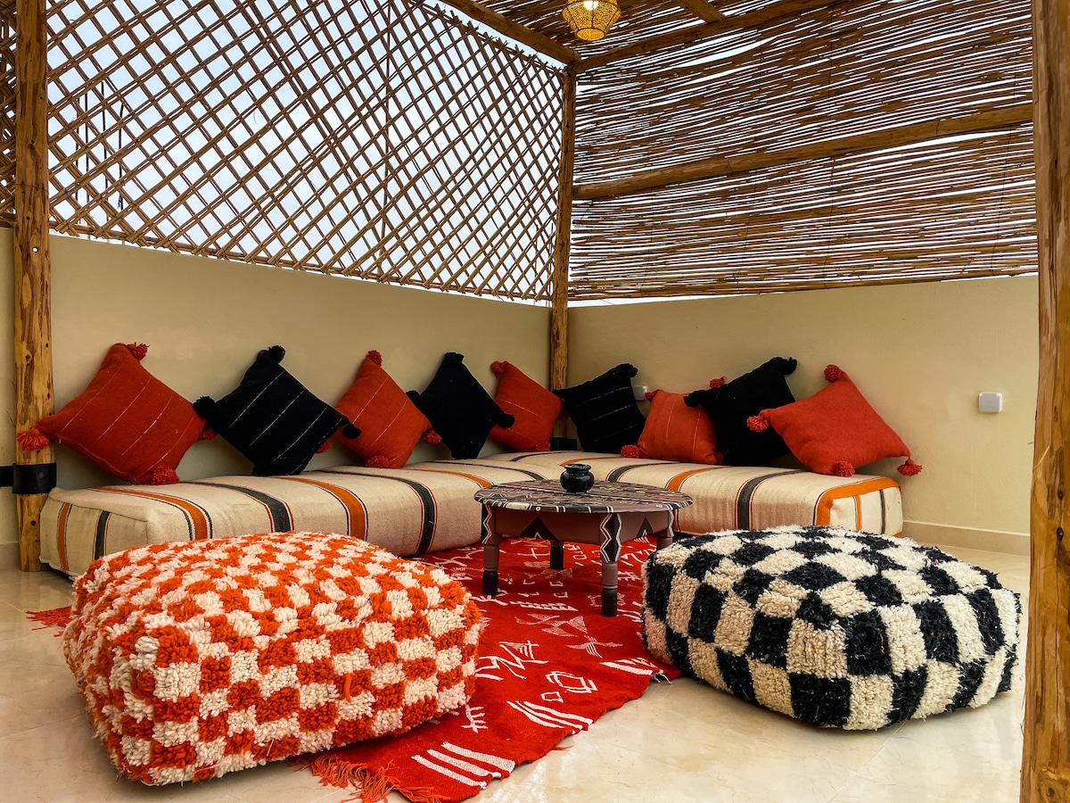 A shaded seating area is presented with colorful, plush cushions arranged around a low table. The space features woven bamboo ceilings and is enhanced by a patterned rug beneath. Two large poufs in contrasting colors add comfort to the environment.