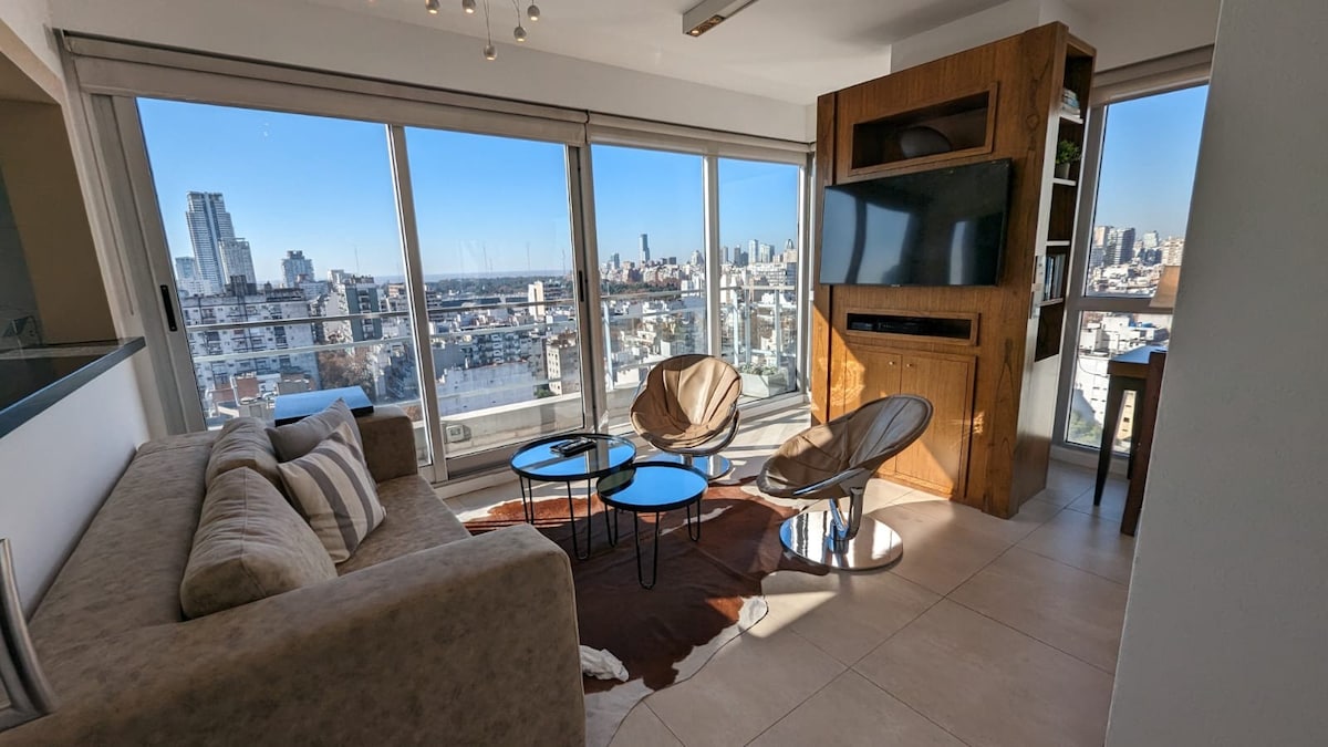 A bright living area features large windows offering panoramic views of the city skyline. Two lounge chairs and a set of coffee tables are positioned on a light-colored rug. A modern entertainment unit houses a flat-screen television, while a comfortable sofa sits invitingly nearby.