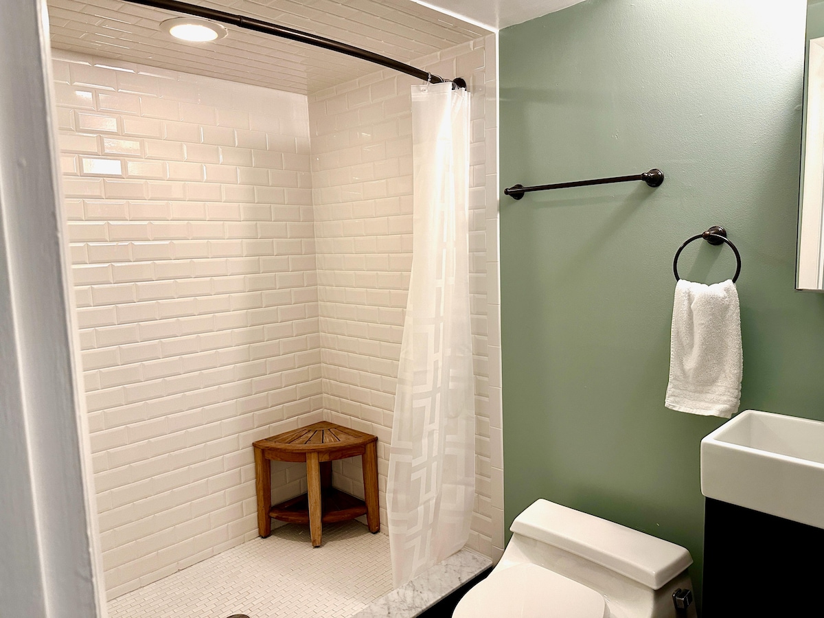 A bathroom features white tiled walls and a spacious shower area, equipped with a clear shower curtain and a wooden bench. A modern toilet is positioned nearby, and a towel hangs on a metal bar. A green accent wall adds a subtle touch.
