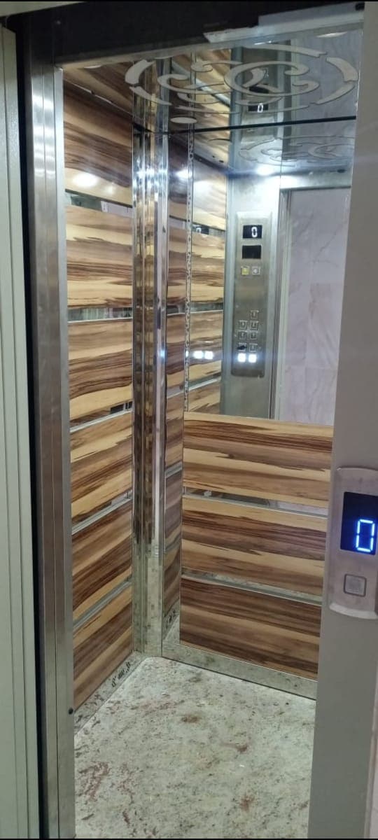 An elevator cabin features sleek, polished wooden paneling and modern metallic accents. The interior is well-lit, showcasing a control panel with buttons for various floors. The spacious dimensions provide ample room for comfortable entry and exit.