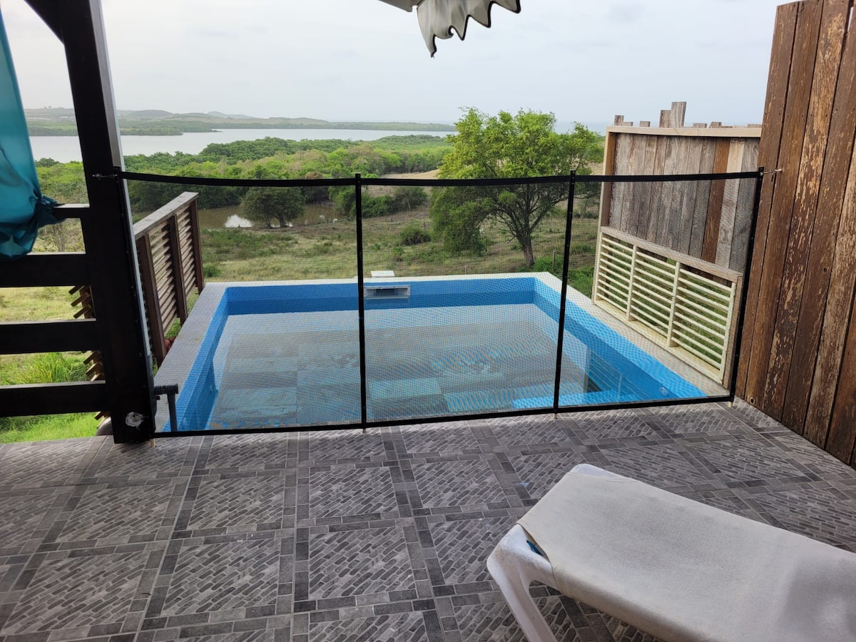 A private swimming pool is viewed from a covered terrace, framed by a safety fence. The pool's clear water reflects the surrounding landscape, showcasing lush greenery and distant water views. A lounge chair rests nearby on the textured stone floor.