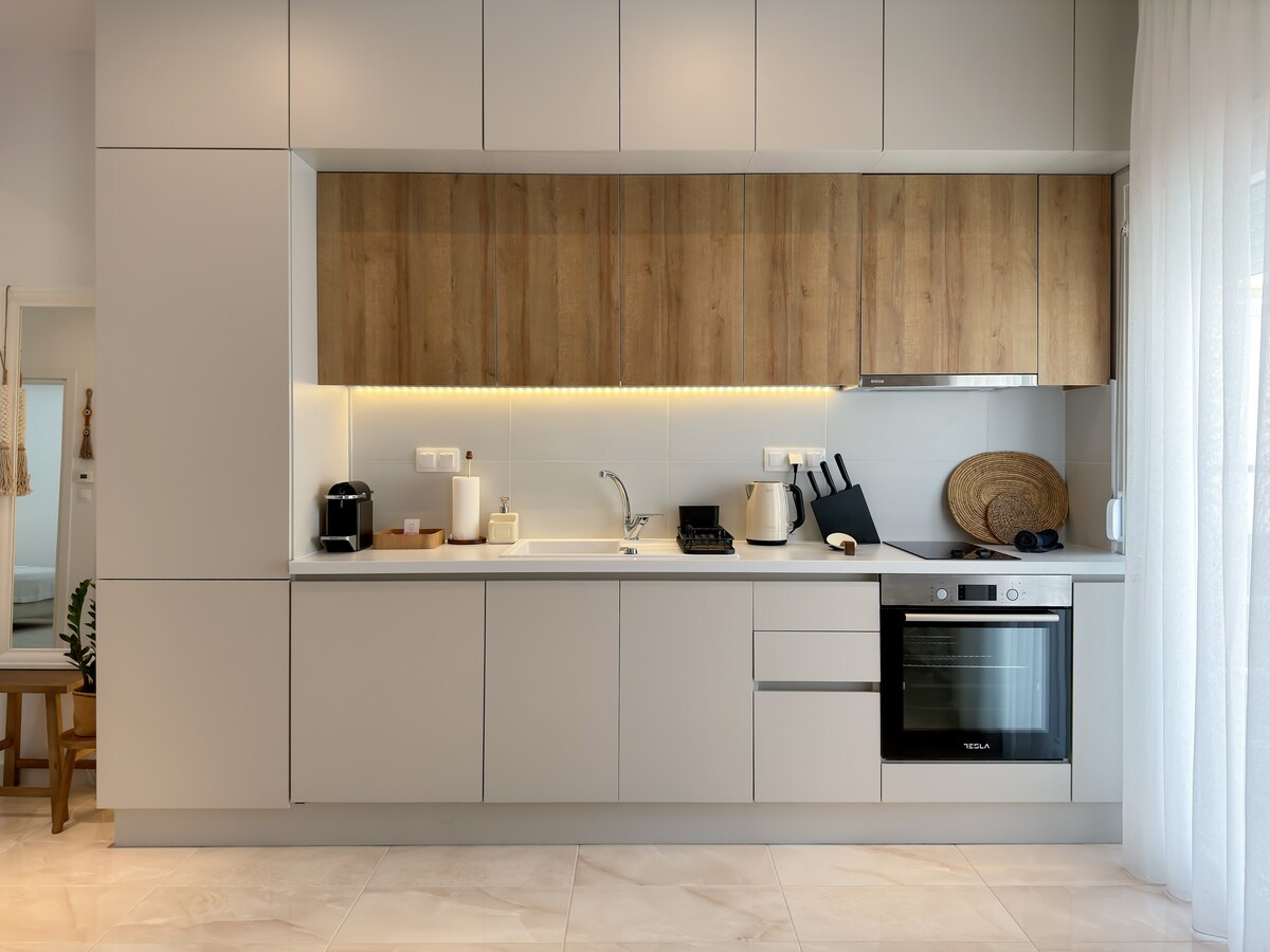 The kitchen area features modern cabinetry in a neutral palette, complemented by wooden accents. Essential appliances include a built-in oven, kettle, and coffee maker. Soft lighting enhances the spacious countertop, while the minimalist design creates an unobtrusive cooking environment.