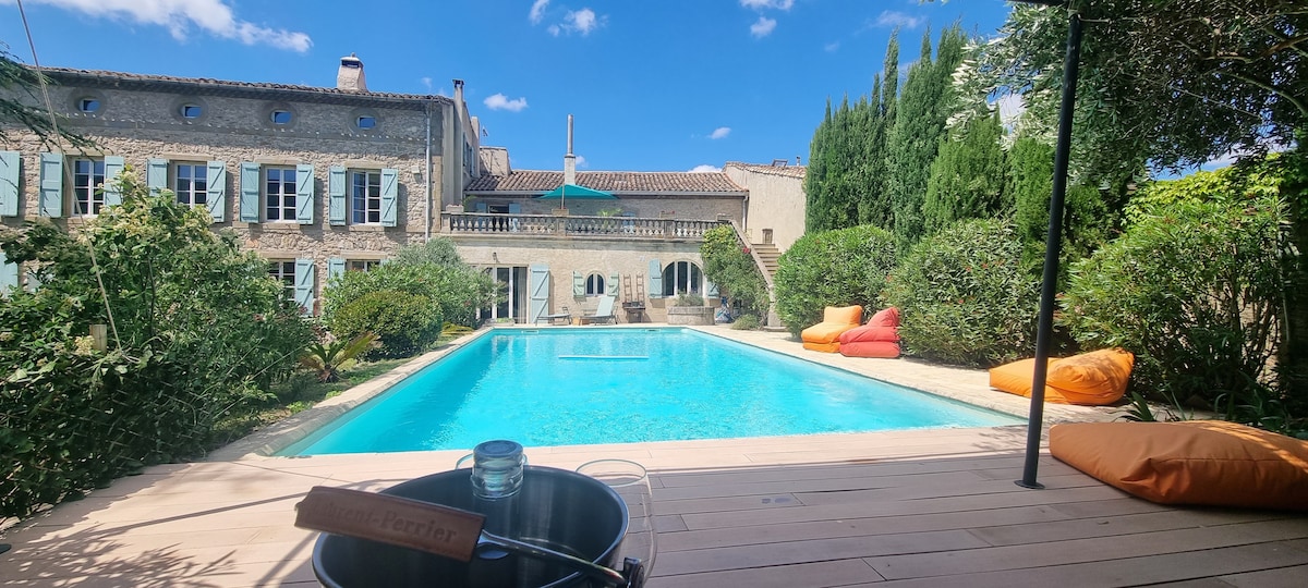 A tranquil swimming pool is surrounded by lush greenery and colorful bean bags, creating a relaxed atmosphere. A spacious stone patio leads to the inviting pool, while the historic house features charming windows and outdoor seating, enhancing the connection between indoor and outdoor living.