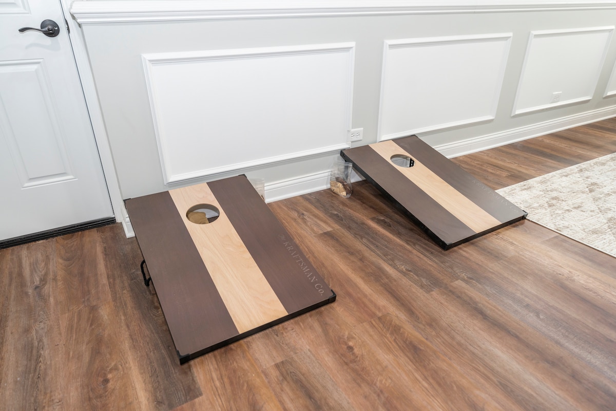 Two cornhole boards are positioned on a hardwood floor, featuring contrasting wood finishes with light and dark stripes. Each board has a circular hole at the top center, providing an opportunity for friendly competition in the game's casual setting.