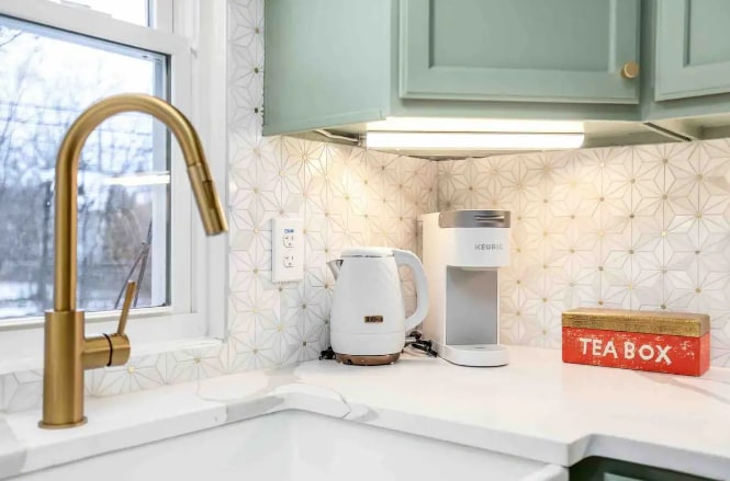 A modern kitchen corner is highlighted featuring a sleek gold faucet and a white kettle beside a coffee maker. Decorative white tiles with geometric patterns adorn the wall, while a red tea box adds a vibrant touch to the countertop.