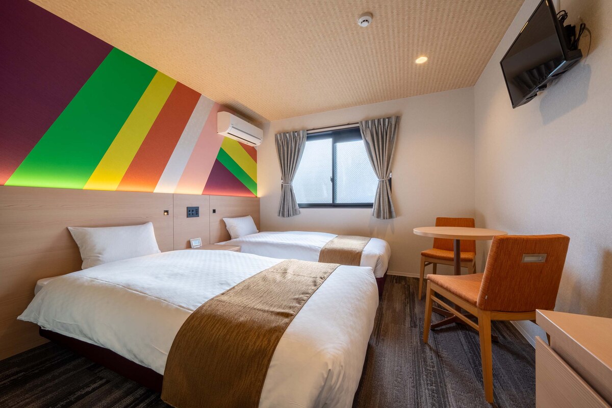 A twin room is presented with two spacious beds featuring contrasting patterned throws. A small table with two chairs is positioned near the window, which is complemented by soft curtains. A colorful accent wall enhances the room's cheerful ambience, while a wall-mounted television is also visible.