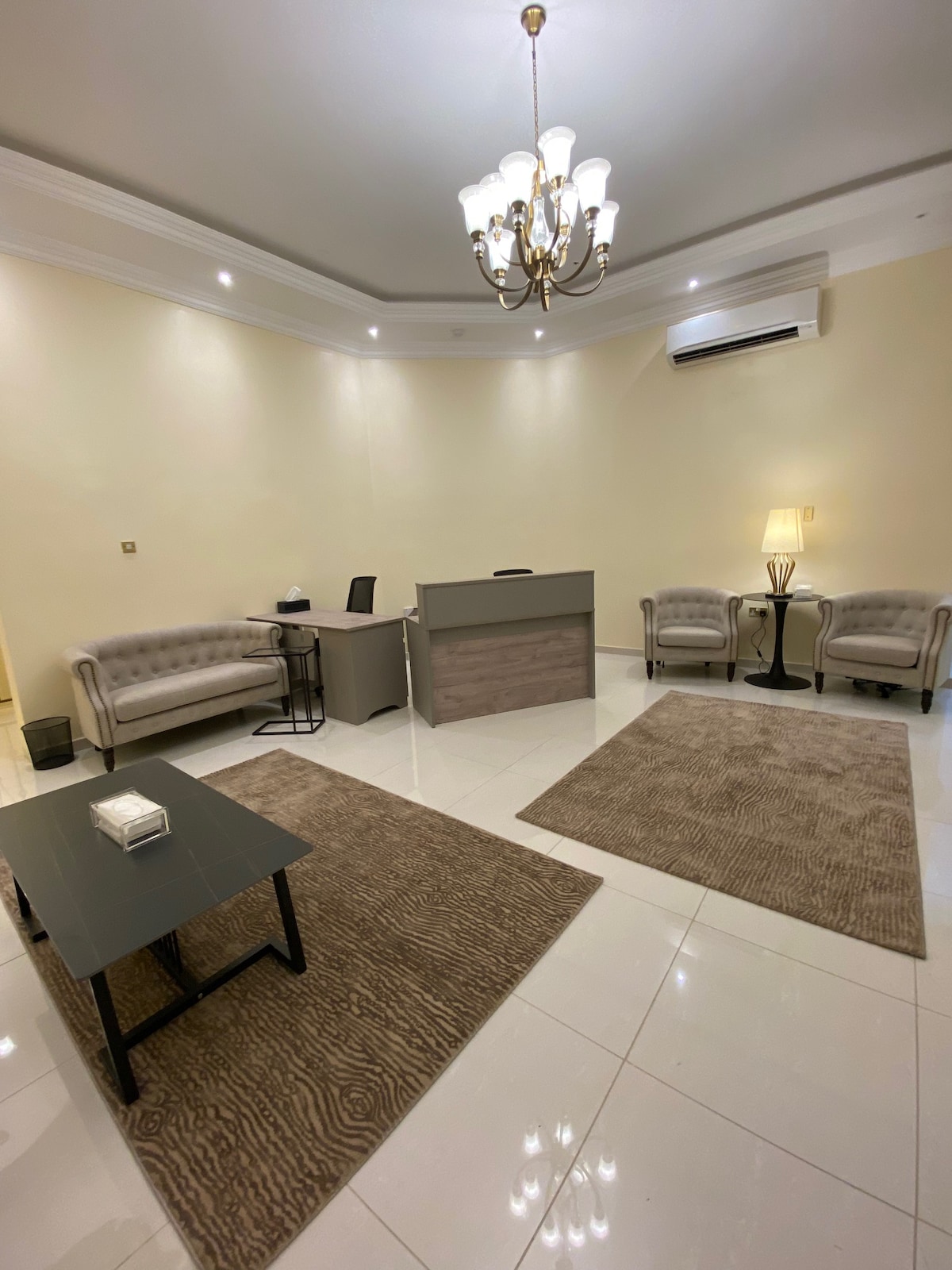 A well-lit office space is presented, featuring a stylish desk with modern chairs. Two plush armchairs are arranged thoughtfully in the seating area, complemented by a sleek side table. Soft rugs add warmth to the polished flooring, while a chandelier provides overhead illumination.