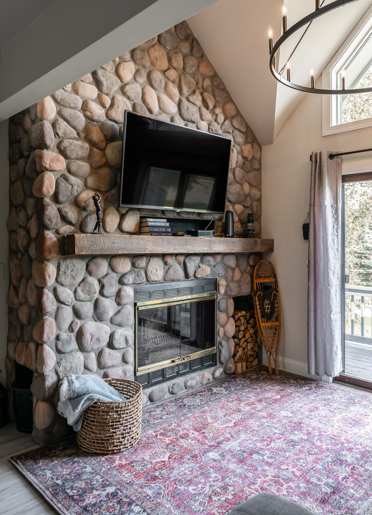 A rustic stone fireplace serves as a focal point, featuring a flat-screen TV mounted above. A wooden mantel displays decorative items, while a basket and a woven log holder provide cozy accents. Soft light filters through nearby windows, illuminating a patterned area rug below.