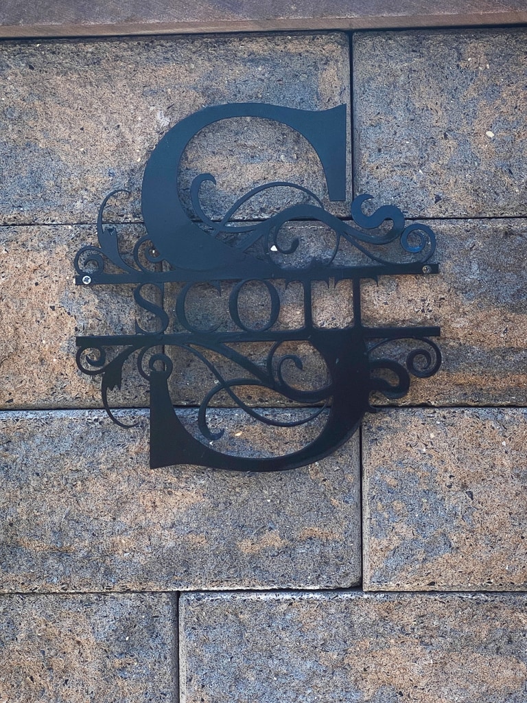 A decorative metal sign hangs against a textured stone wall. The sign features the name 'Scott' in an elegant font, with intricate flourishes surrounding the letters, providing a distinctive welcome to guests.