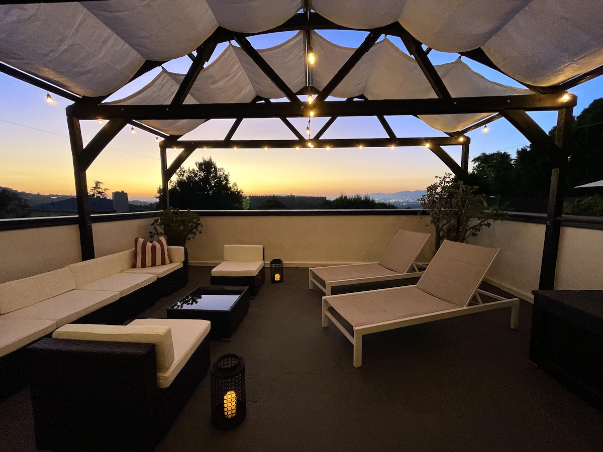 An outdoor patio is featured under a canopy with soft lighting, creating a cozy ambiance. Two sun loungers are positioned beside a sectional seating area. A low table sits at the center, all surrounded by scenic views of the hills and the sunset.