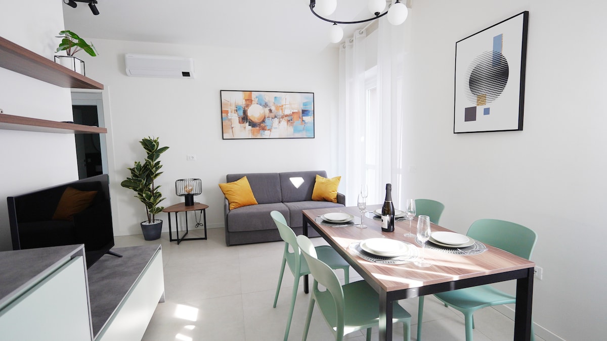 The living area features a sofa bed adorned with bright yellow cushions, paired with a sleek dining table set for four. A decorative plant adds a touch of greenery, while a television is positioned nearby. Large windows allow natural light to fill the space.